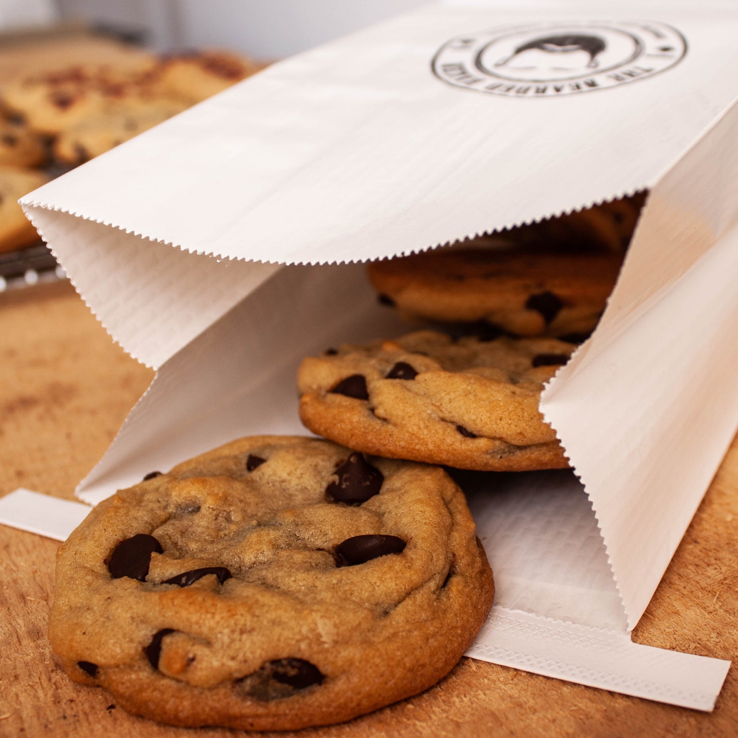 M.O.M. Chocolate chip cookies from the Bearded Baker spilling out of a paper bag on a wooden surface